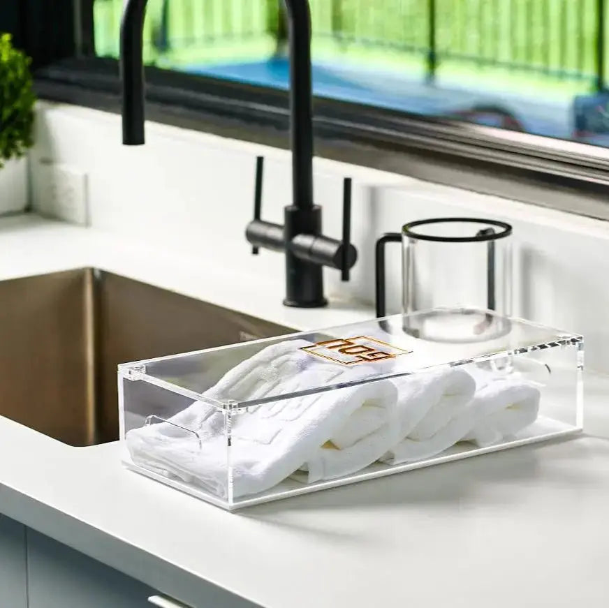 Clear lucite Pesach towel box with folded white towels and black faucet displayed on white kitchen counter near sink.