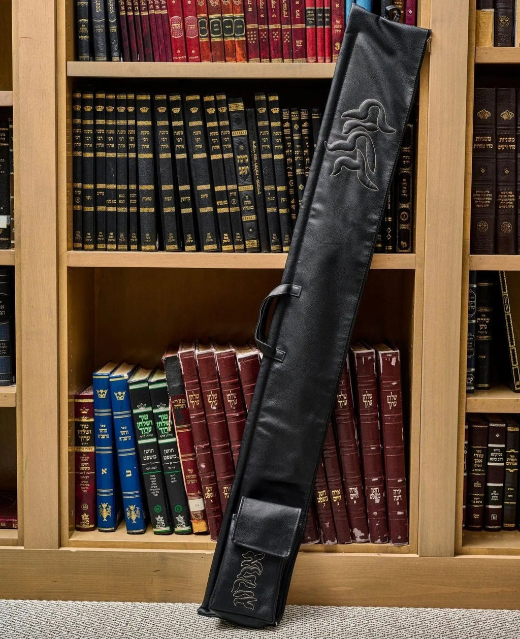 Black leather Pillow Script lulav case displayed standing upright in wooden bookshelf, showing embossed design and carrying strap for Sukkot holiday.