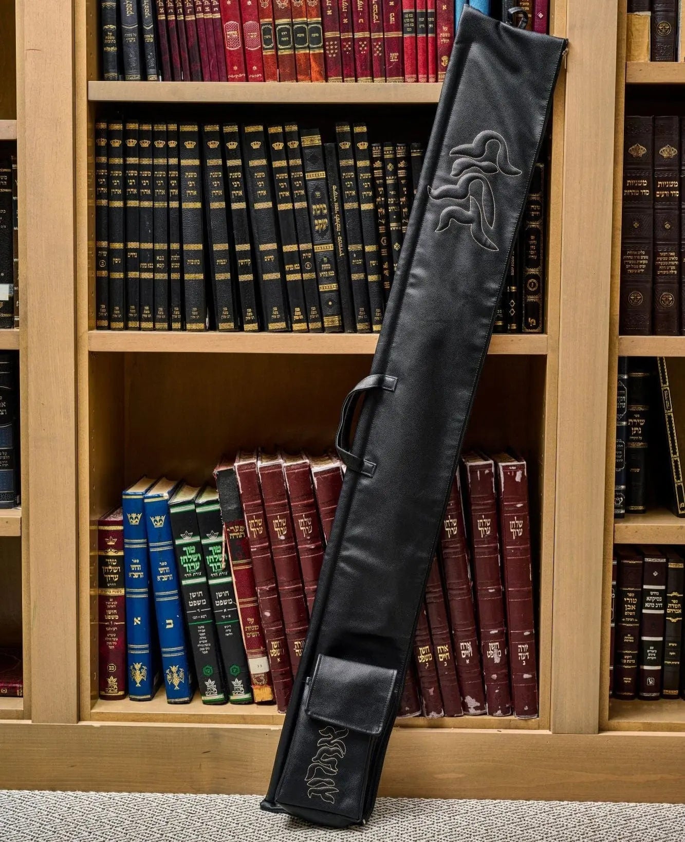 Black leather Pillow Script lulav case displayed standing upright in wooden bookshelf, showing embossed design and carrying strap for Sukkot holiday.