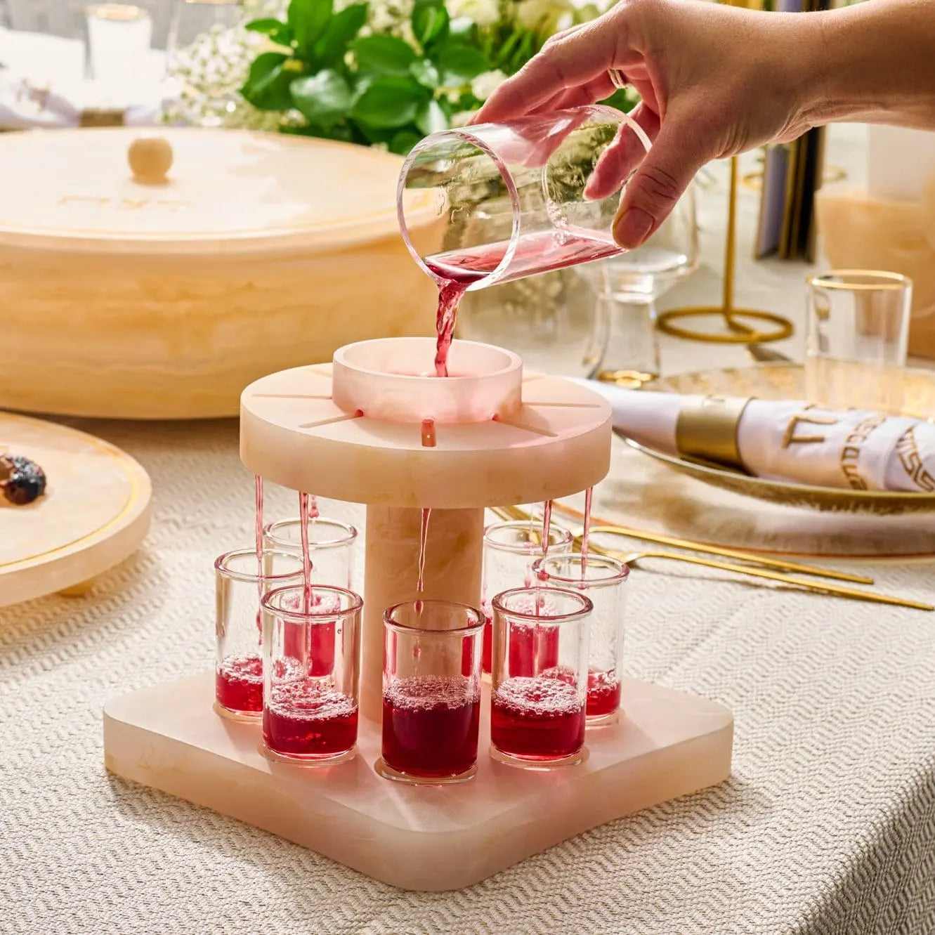 Wooden resin Kiddush fountain with pink glass cups arranged on tiered wooden base, displayed on kitchen counter with Shabbat items.