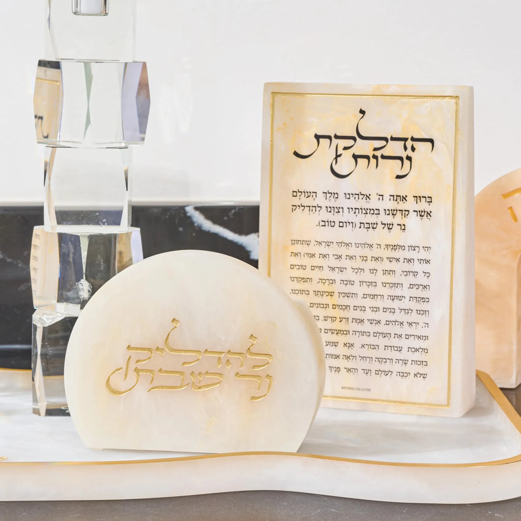 A small cream-colored  resin plaque with gold Hebrew lettering sits in front of a matching resin matchbo displayed on a white marble surface 