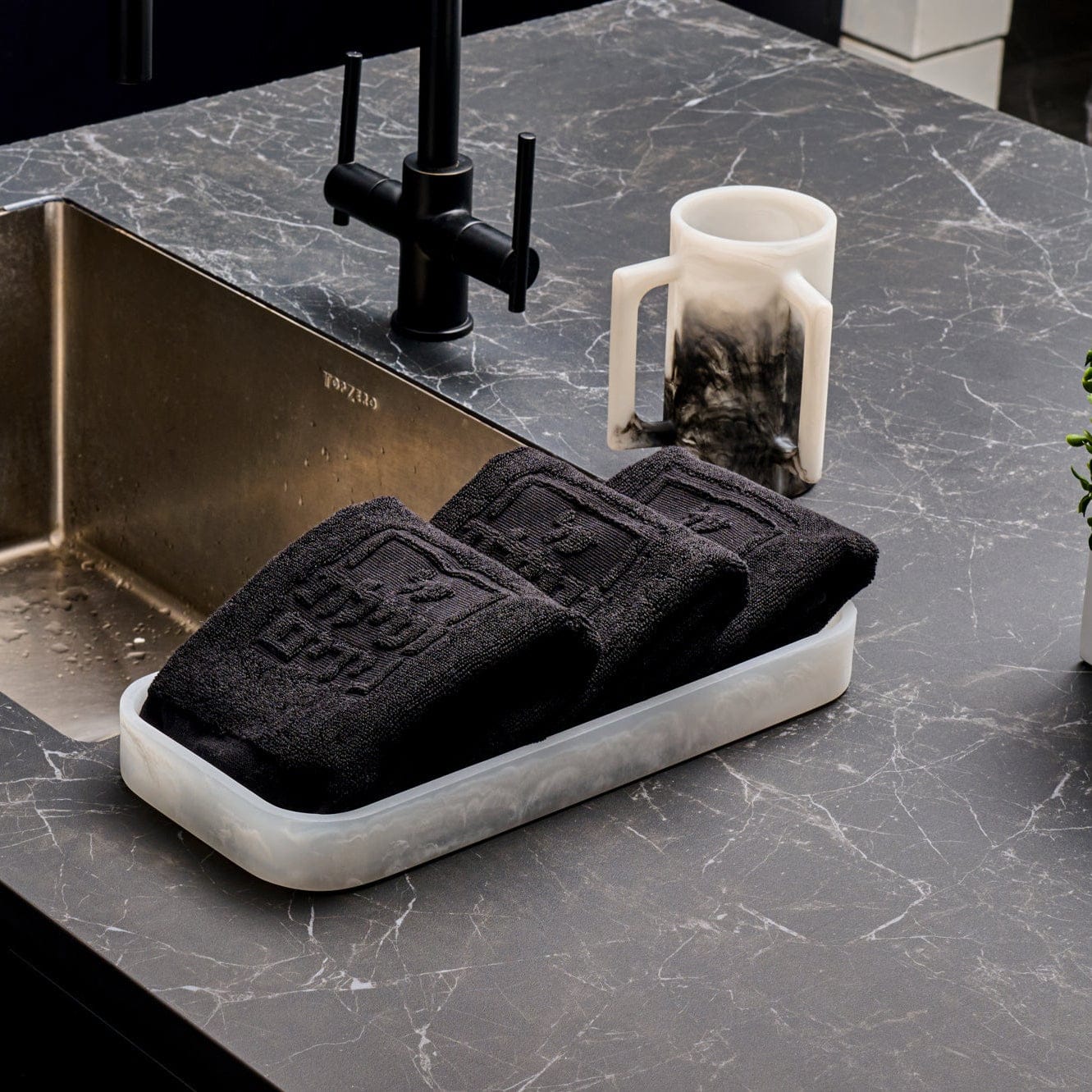 Rectangular marble-look resin tray holding folded black embossed towels, styled on a dark stone countertop beside a modern matte black faucet, stainless sink, and black-and-white resin washing cup.
