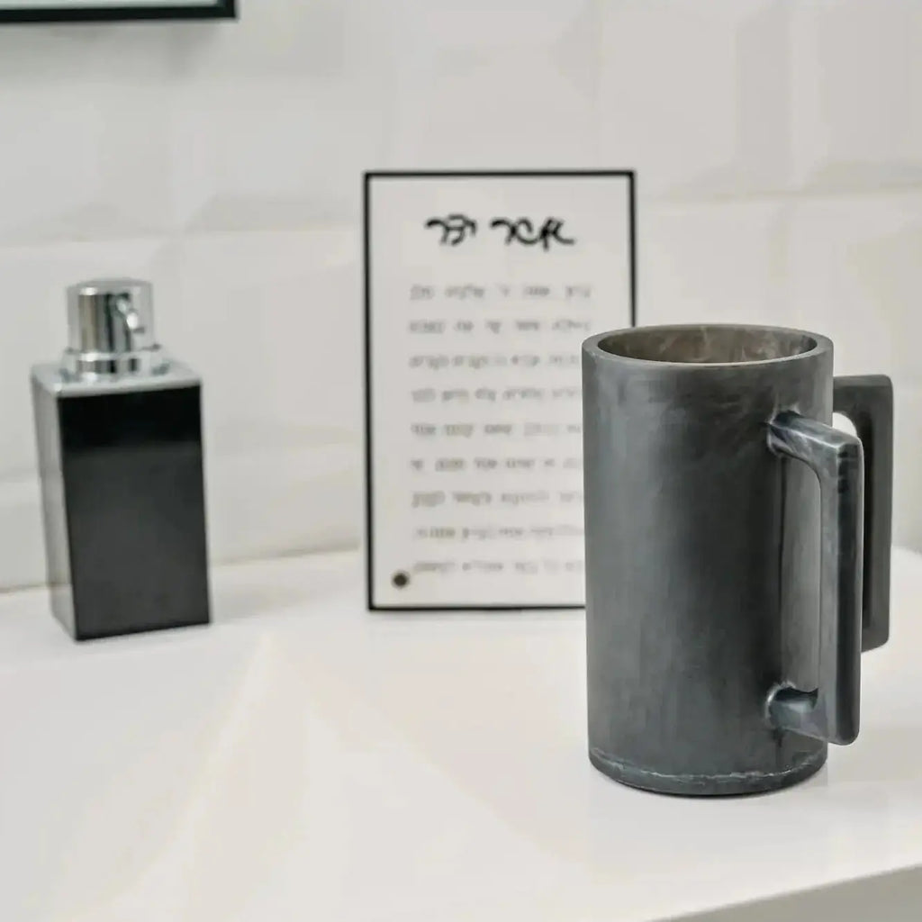 Black resin washing cup displayed on white surface with matching black bottle and instructional card showing traditional Jewish handwashing ritual guide.
