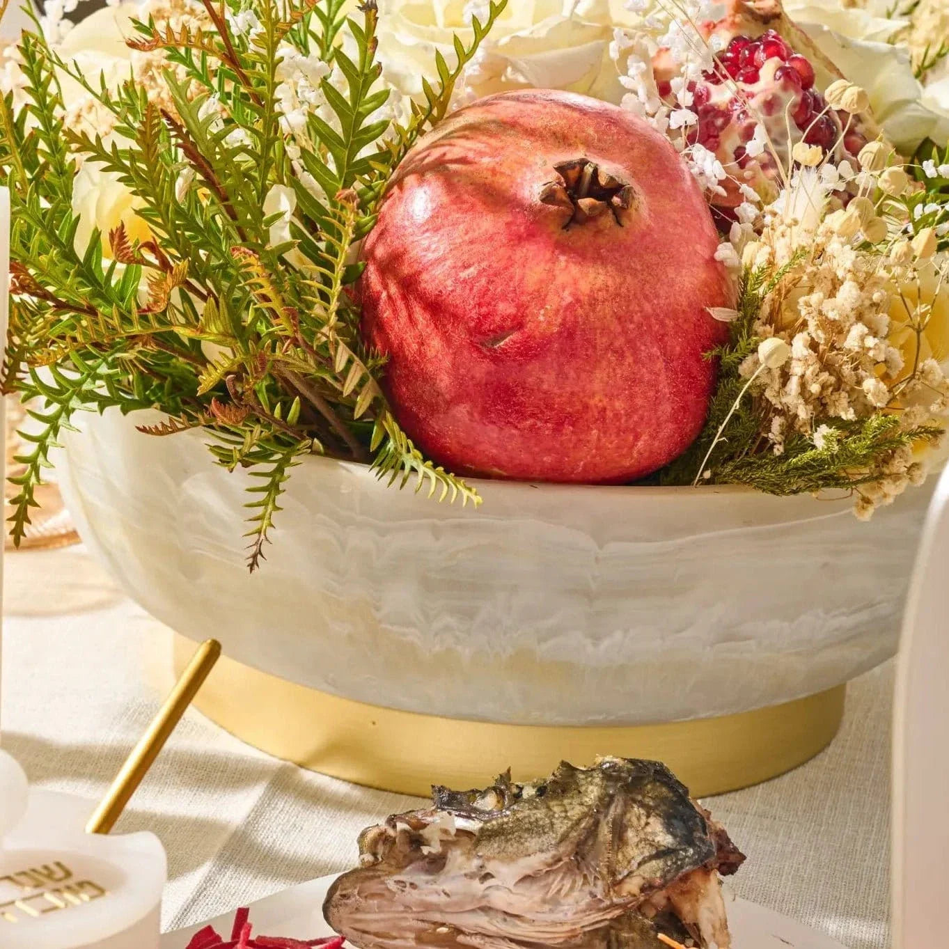 A festive rosh-hashana table setting featuring a white and gold resin bowl filled with greenery, dried flowers, and a whole pomegranate, alongside a printed Hebrew simanim card.