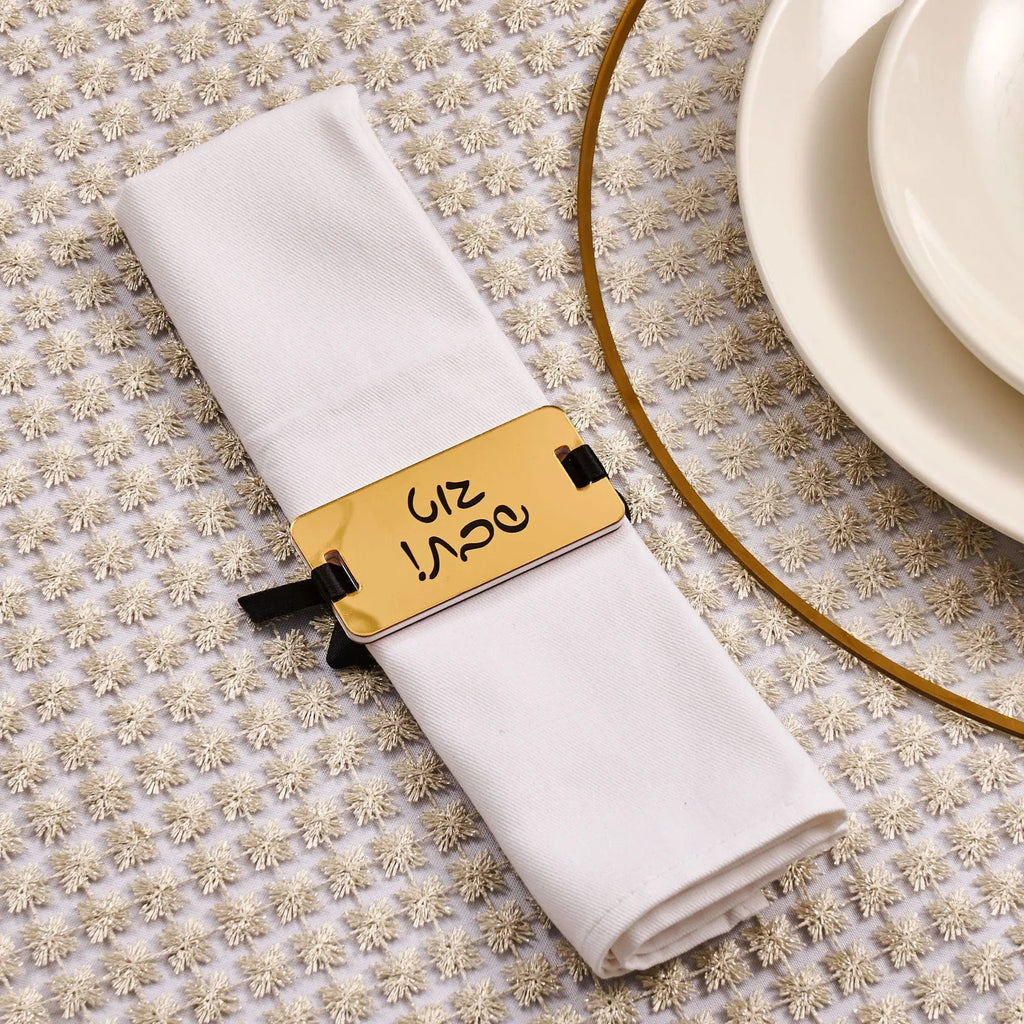 White Shabbos napkin wrap with gold metallic band featuring Hebrew text, displayed on folded white napkin next to place settings