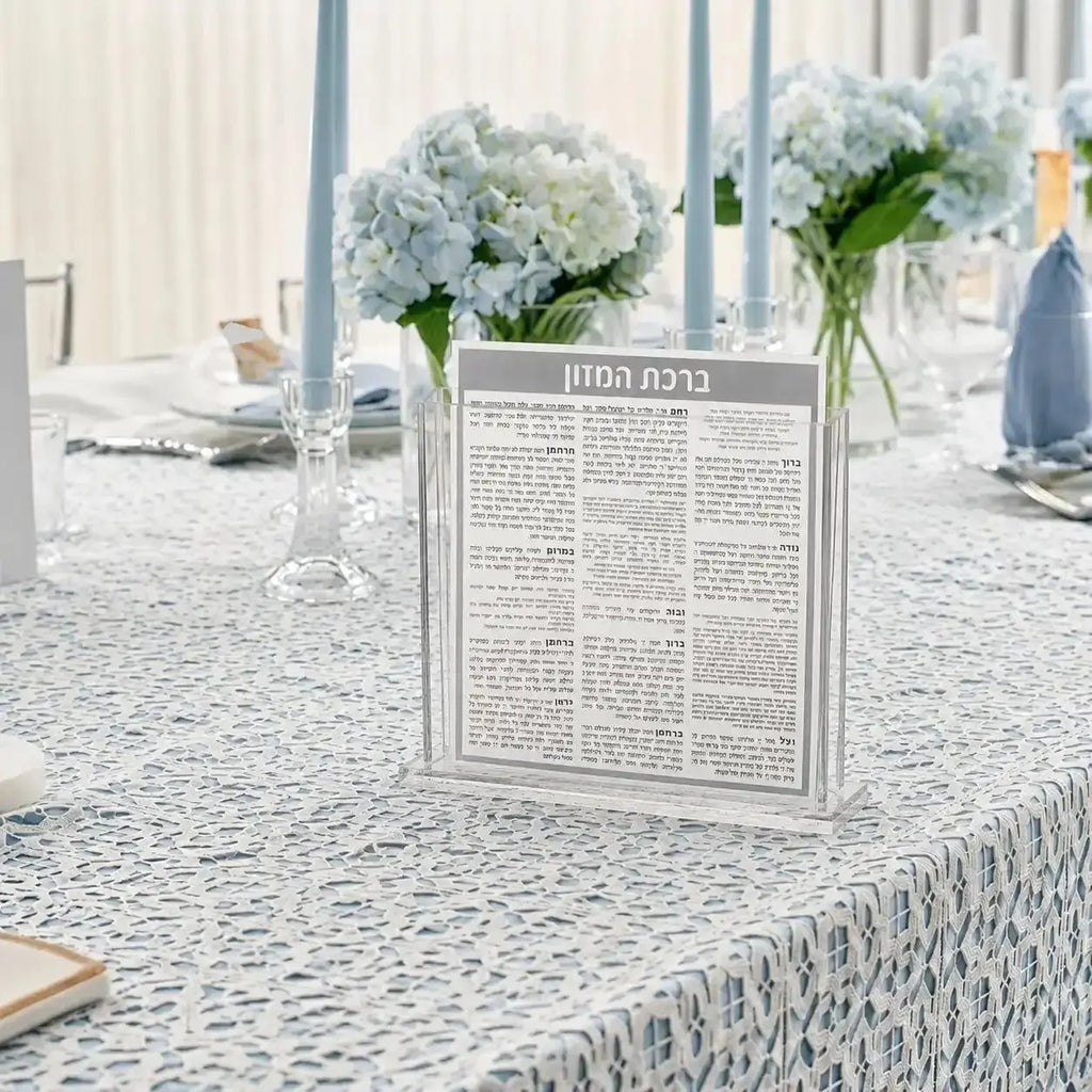Square Bencher Set with white flowers in glass vases on patterned tablecloth, black and silver Jewish table decor for Shabbat.