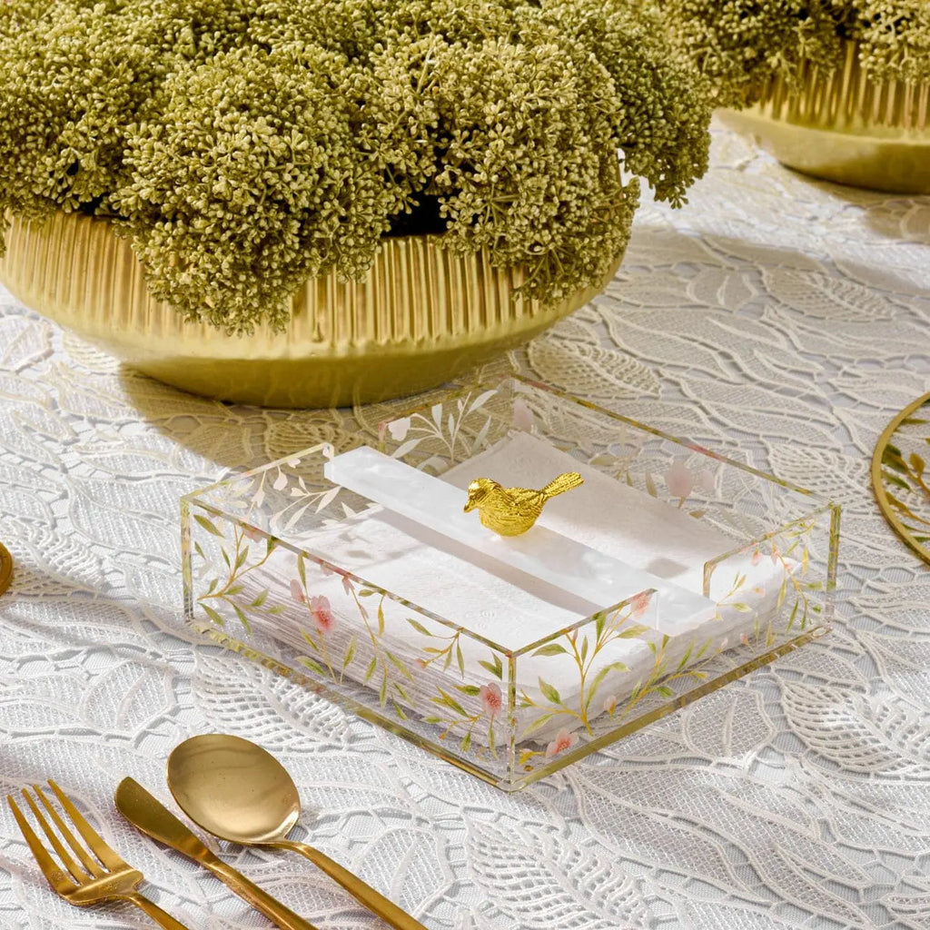Square floral bird napkin holder in clear acrylic with gold metal accents, displayed on a white table with matching gold utensils and tableware.