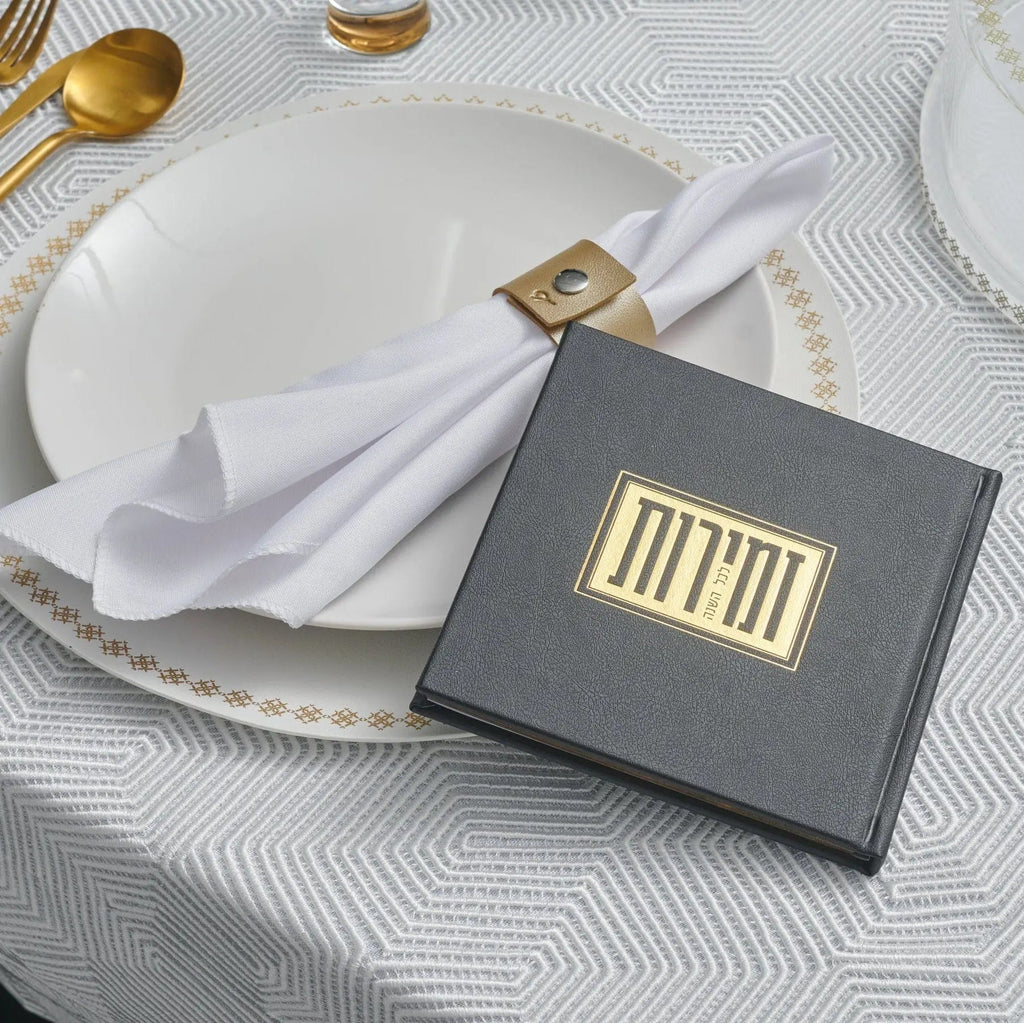 Elegant black and gold square Shabbos bencher set displayed on white plate with gold napkin ring, featuring formal table setting with gold utensils and patterned tablecloth.