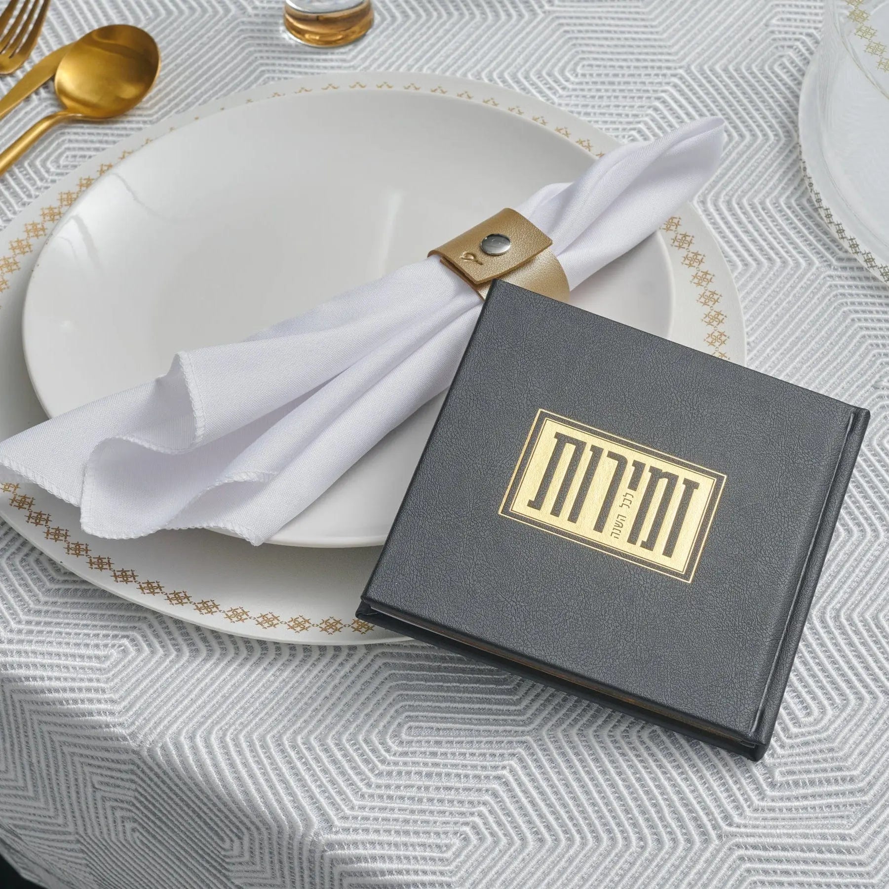 Elegant black and gold square Shabbos bencher set displayed on white plate with gold napkin ring, featuring formal table setting with gold utensils and patterned tablecloth.