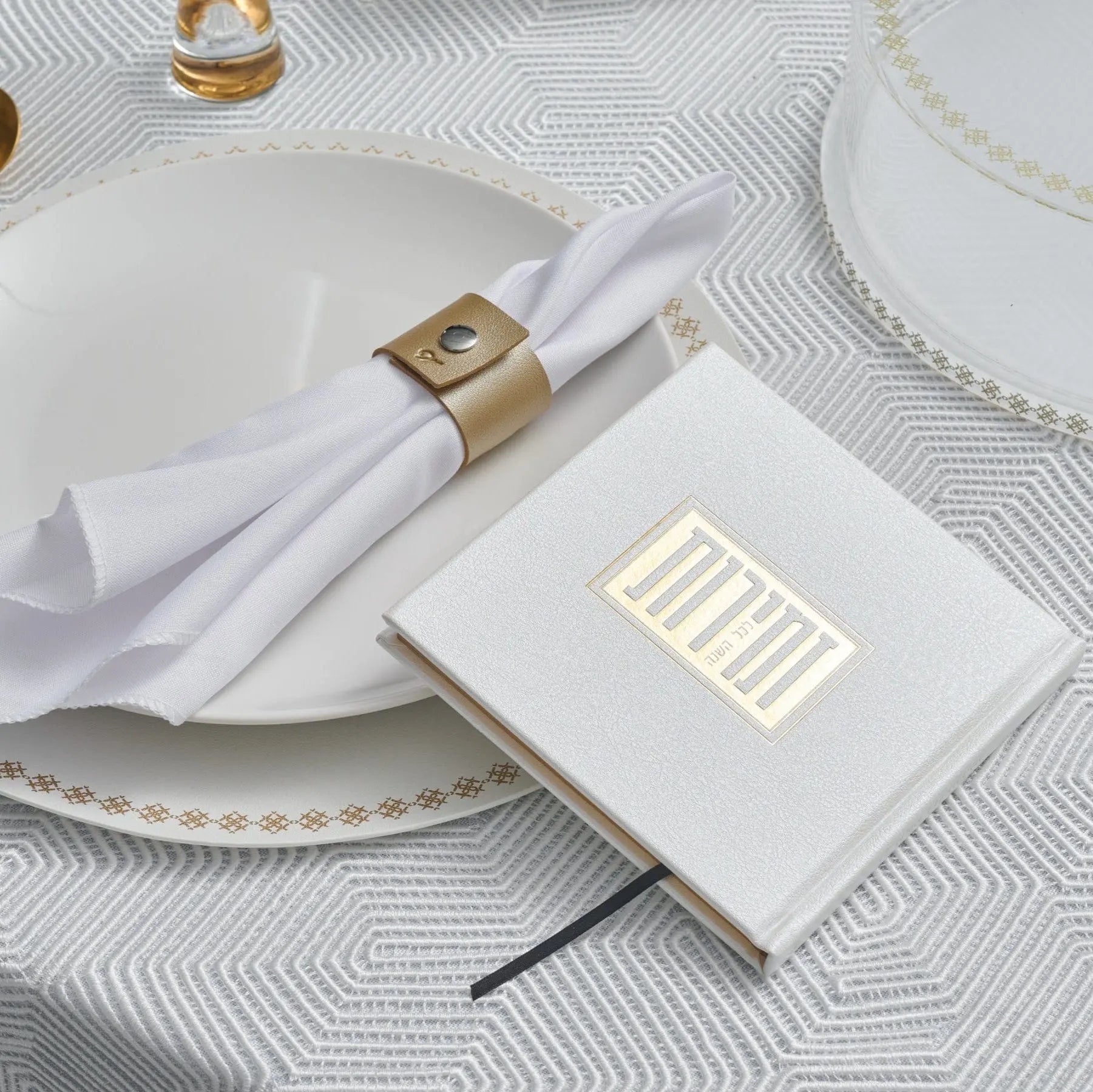 Elegant white Shabbos bencher with gold accents displayed on a place setting with white napkin and gold napkin ring, perfect for holiday entertaining.