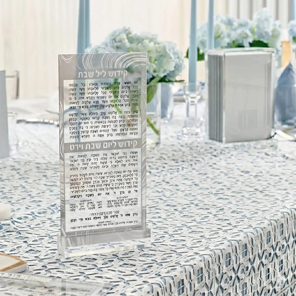 Silver Swirl Kiddush Card in lucite displayed on a patterned tablecloth with white flowers and place settings in background