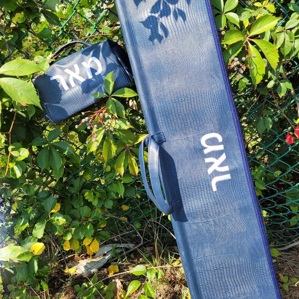 Blue textured leather esrog bag from Waterdale Collection, displayed outdoors on green foliage with carrying strap visible.