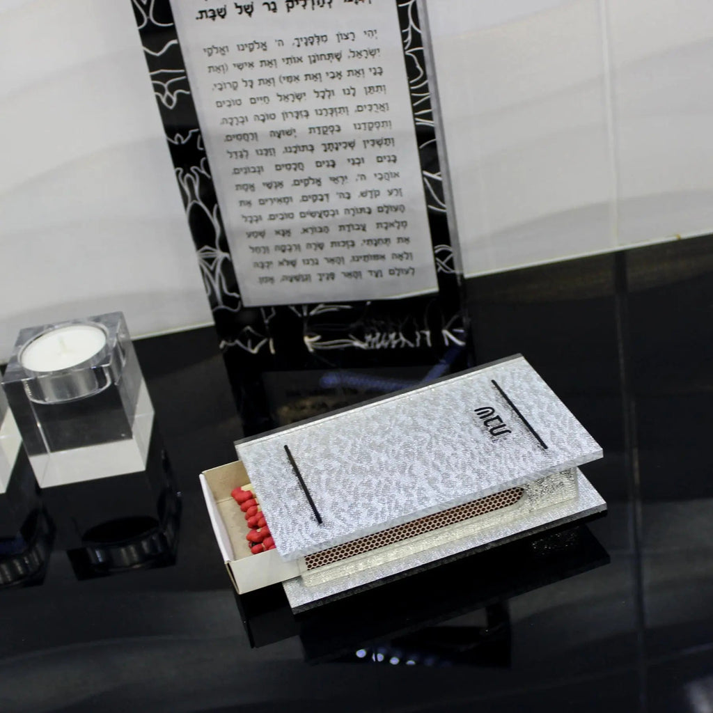 Silver shimmer matchbox holder with red matches displayed on black surface, paired with white candle and Hebrew text backdrop for Shabbat candle-lighting.