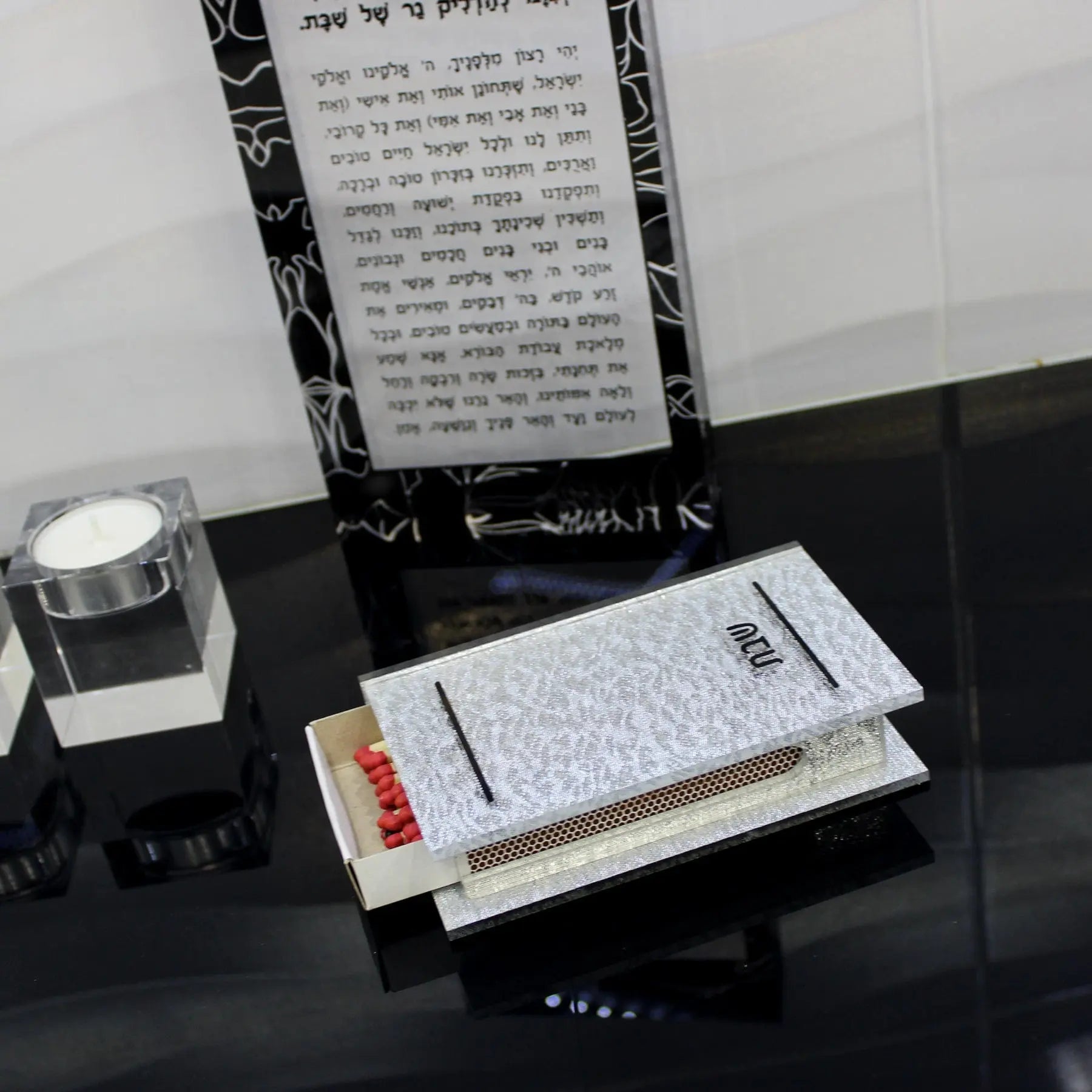 Silver shimmer matchbox holder with red matches displayed on black surface, paired with white candle and Hebrew text backdrop for Shabbat candle-lighting.
